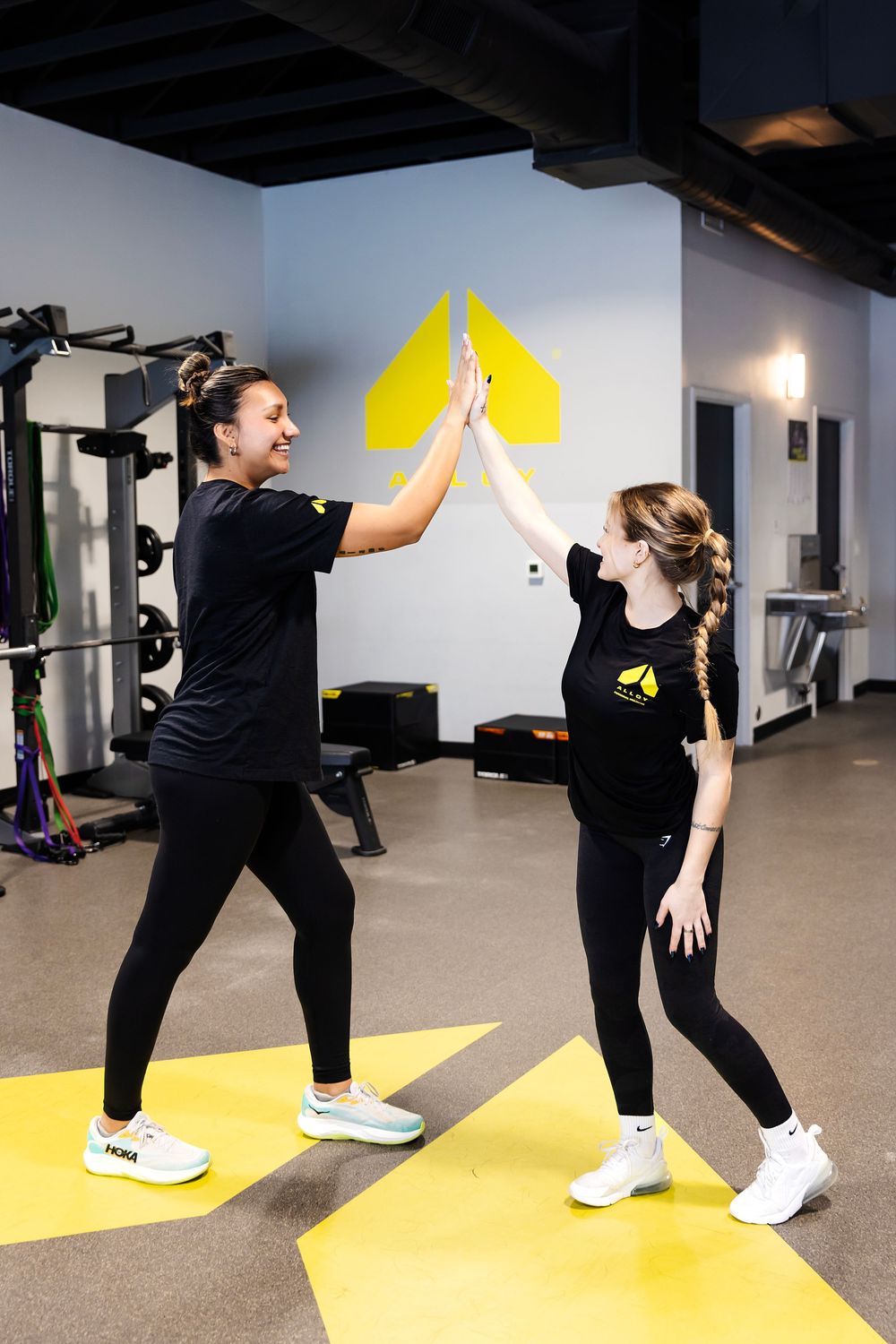 Two women in matching black gym outfits high-fiving in a modern fitness studio with yellow accents.