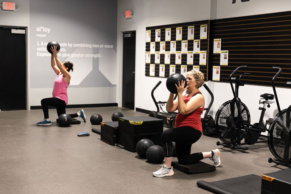 Two women perform weighted lunges in a modern gym, one lifting a medicine ball overhead and the other holding a plate.