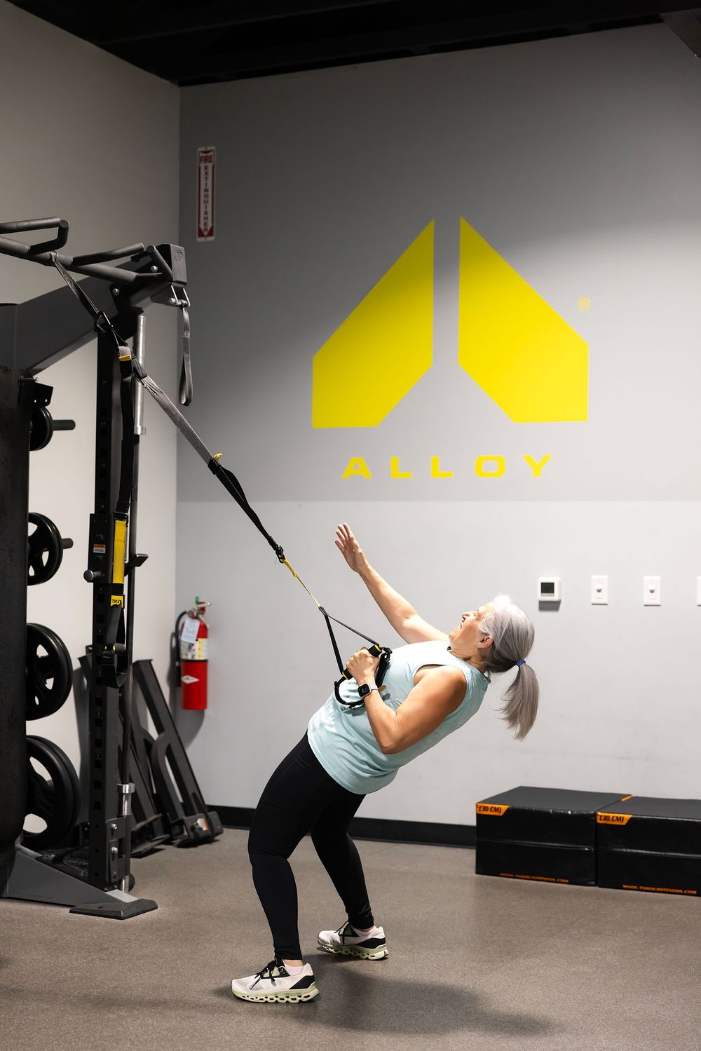 Woman performing TRX suspension training exercise at Alloy fitness gym with yellow logo on wall.
