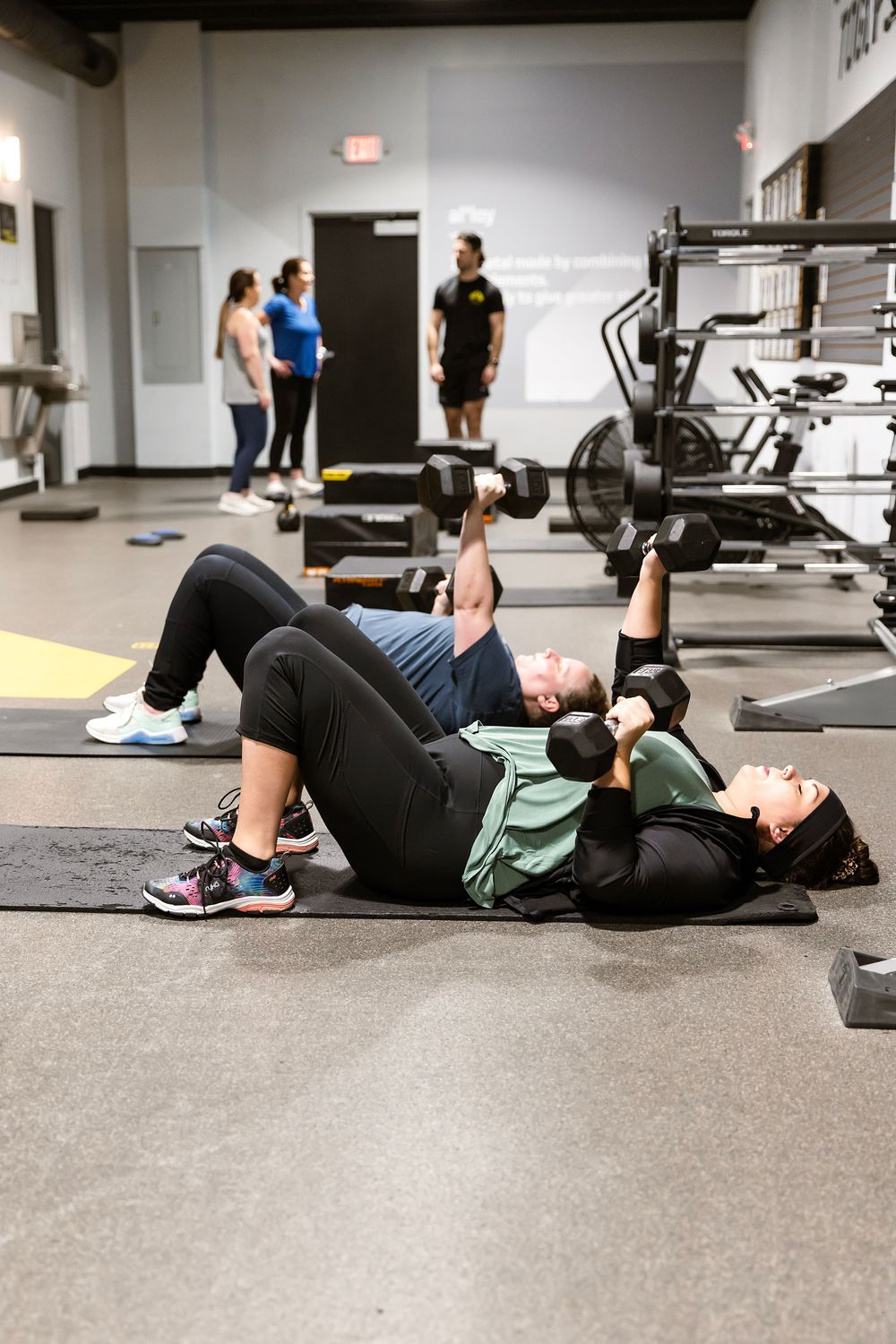 Two people perform dumbbell floor presses on gym mats while others exercise in a modern fitness center.