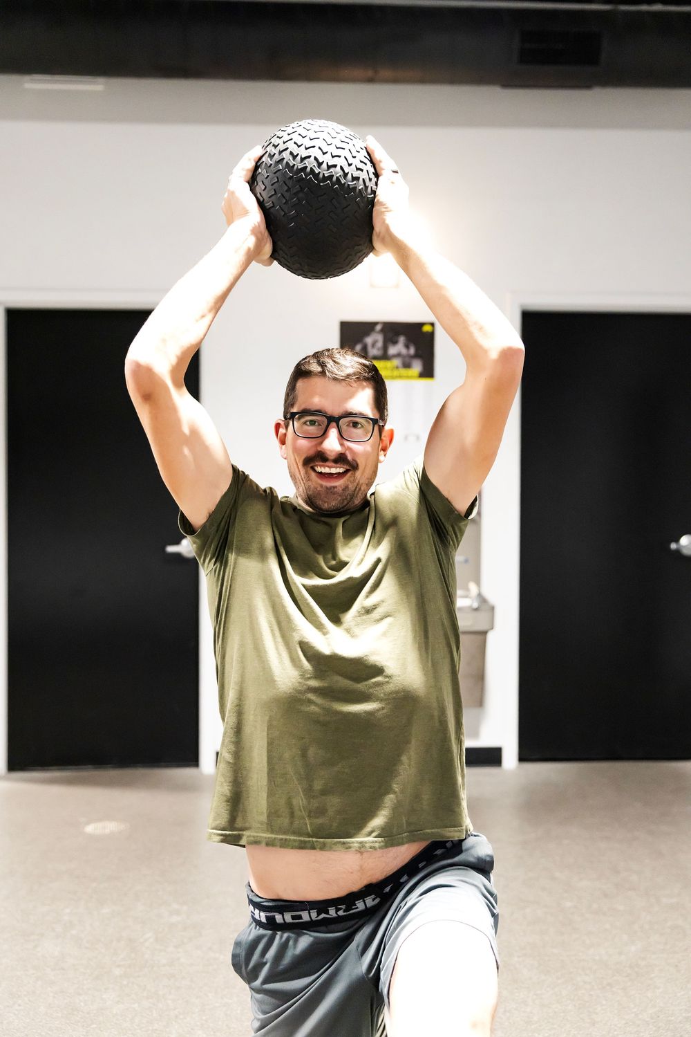 Smiling man in glasses and green t-shirt lifting a black medicine ball overhead during a gym workout session.