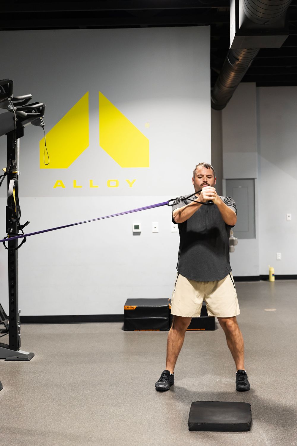 Man performing resistance band exercise at Alloy fitness gym, pulling band in wide stance position on workout floor.