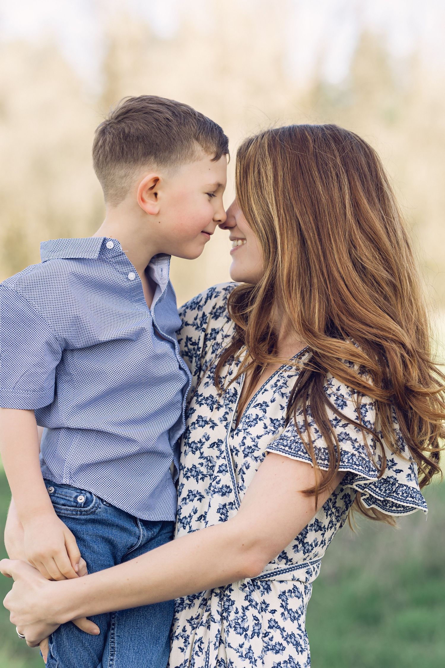 Mother and son touching noses and smiling warmly while embracing outdoors in soft natural light.