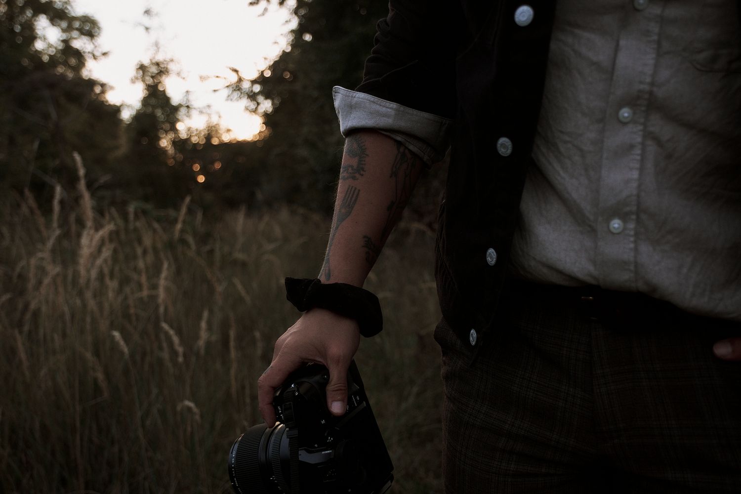 Tattooed photographer holding a camera in a field at dusk, wearing a black jacket over a gray shirt.