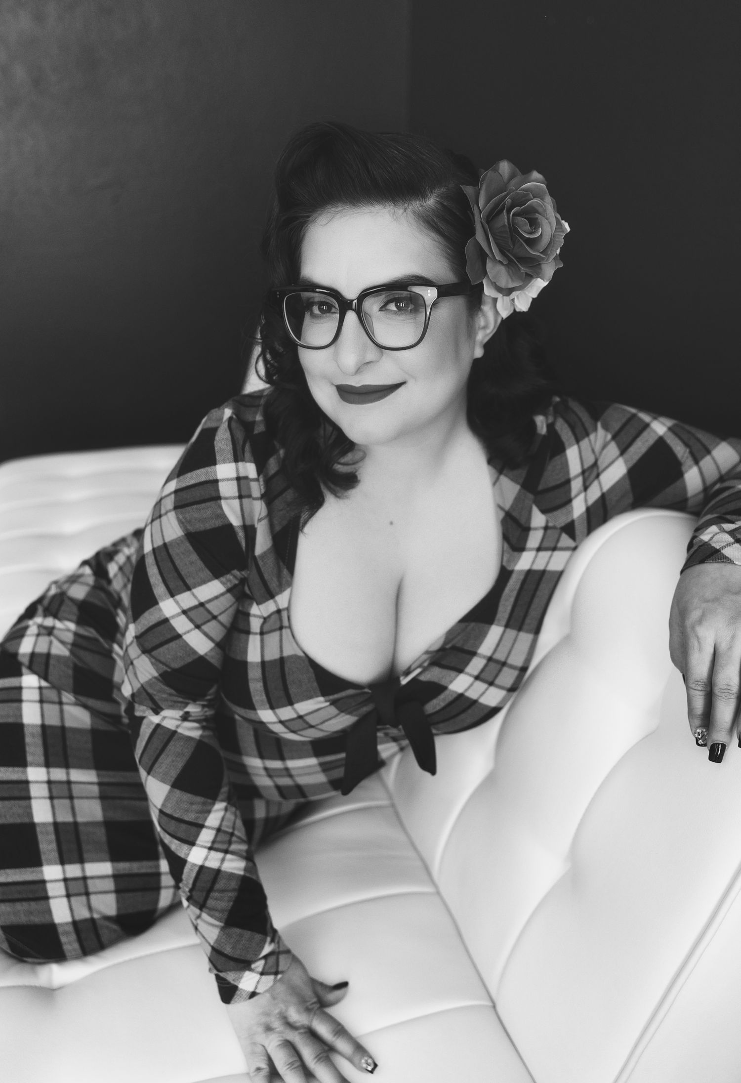 Black and white pin-up style photo of a woman in plaid with glasses and a rose hair accessory, posing on a white couch.