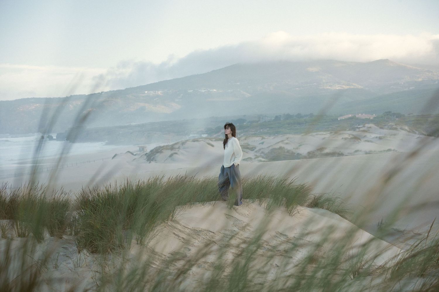 A woman stands alone on sandy dunes with coastal grass, misty mountains, and ocean in the background.