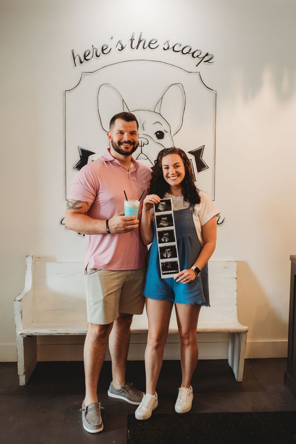 A couple poses against a white wall with decorative text and a church pew style bench.