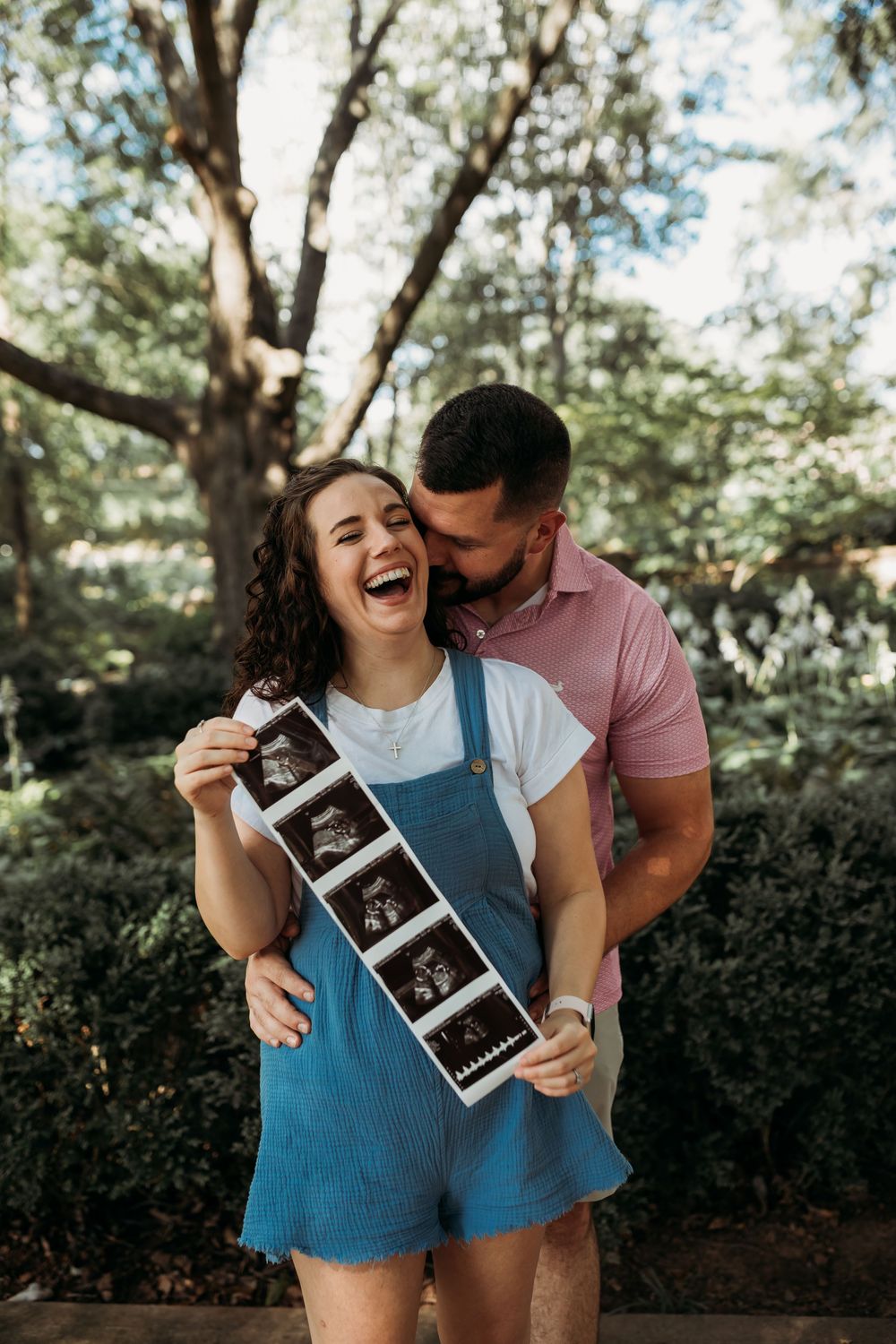 A couple shares a joyful moment outdoors while holding an ultrasound photo in a natural setting.