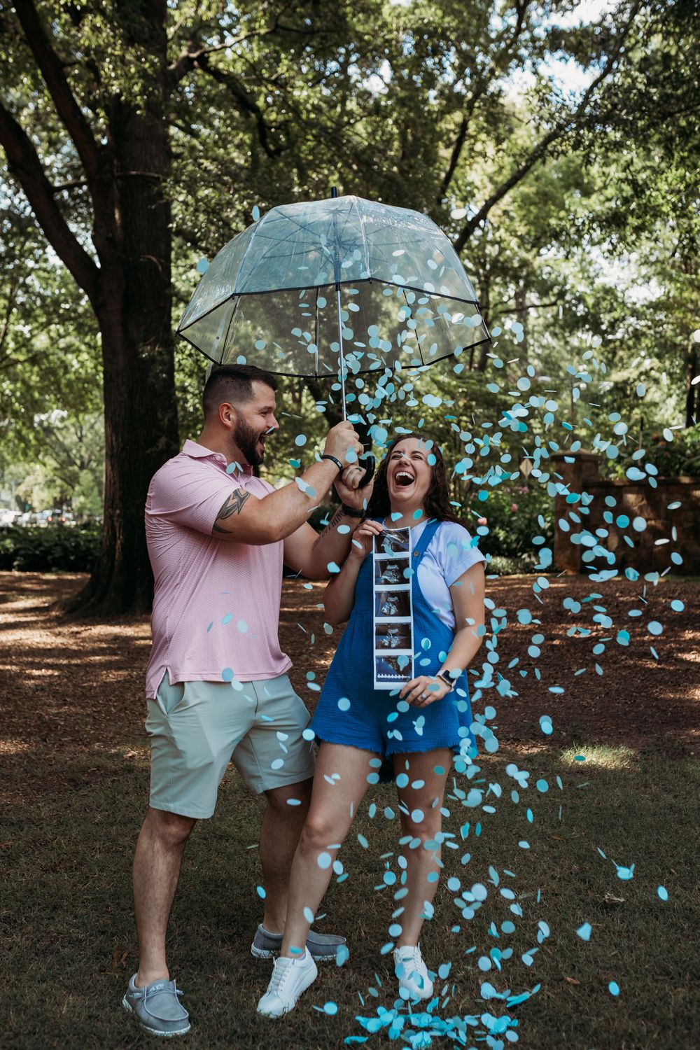 A joyful couple celebrates under a clear umbrella with blue confetti raining down in a wooded setting.