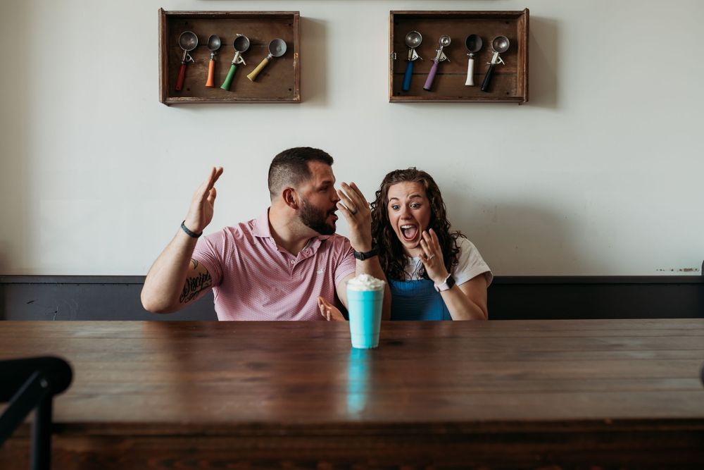 A couple sits at a long wooden table with a turquoise tumbler while engaging in playful conversation.