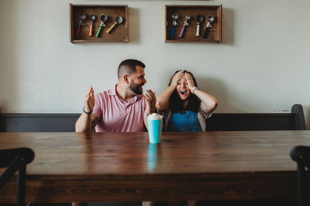 Two people share a lighthearted moment at a minimalist dining table with wall art in the background.