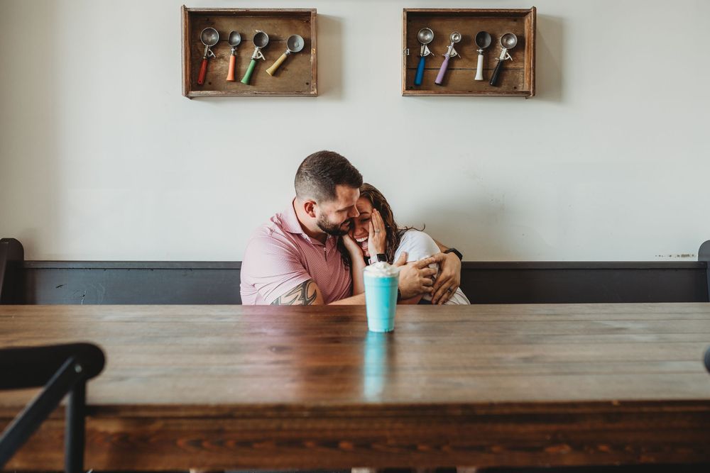 A couple shares a milkshake at a wooden table in a cafe with decorative wall art above.