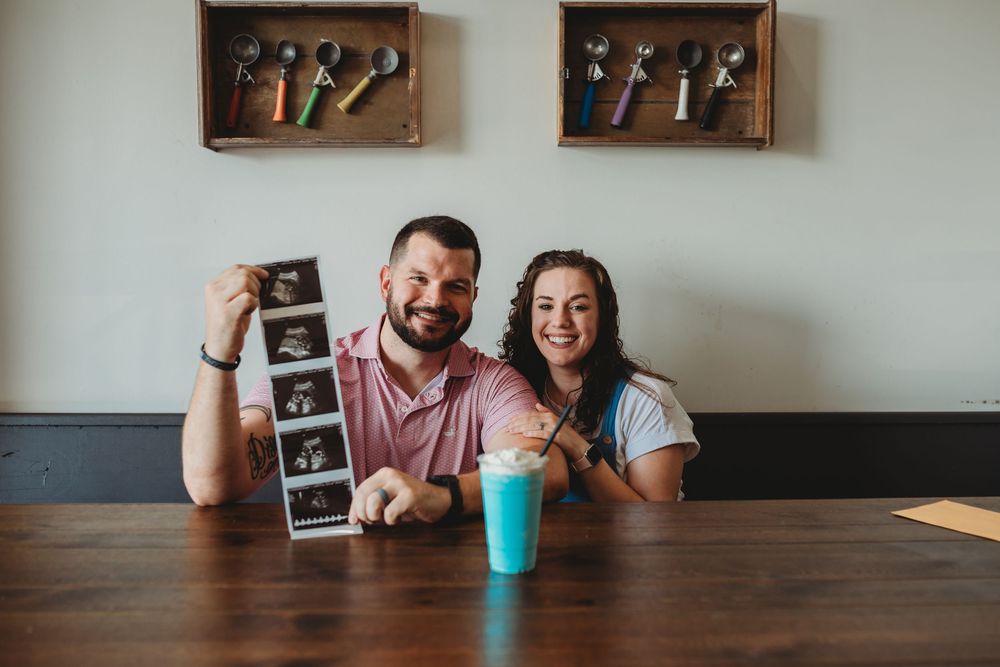 Couple sitting at wooden table holding ultrasound photos while smiling at camera in pregnancy announcement series.