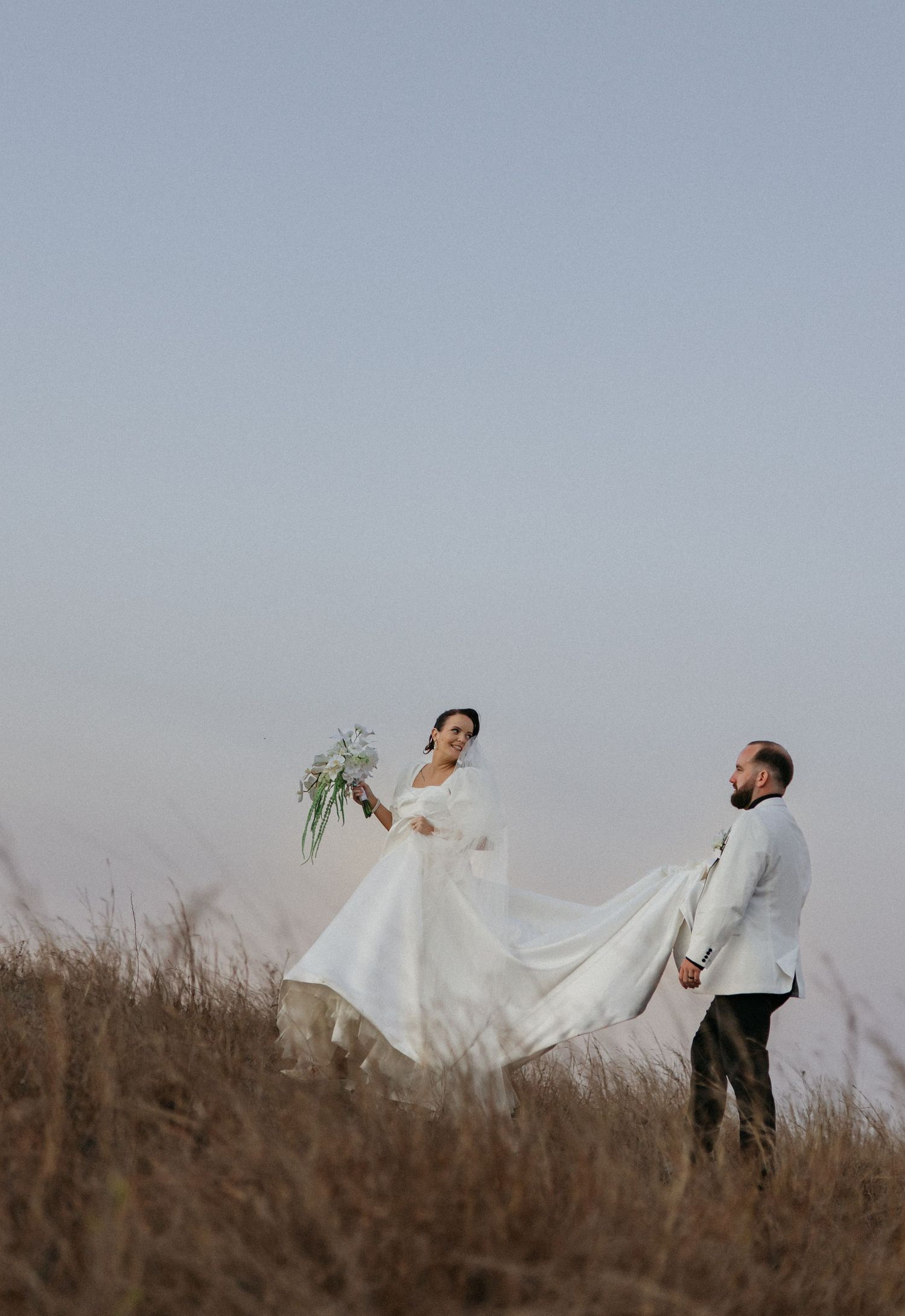 A bride in a flowing white gown holds a bouquet while a groom in a white suit lifts her dress train in a golden field.