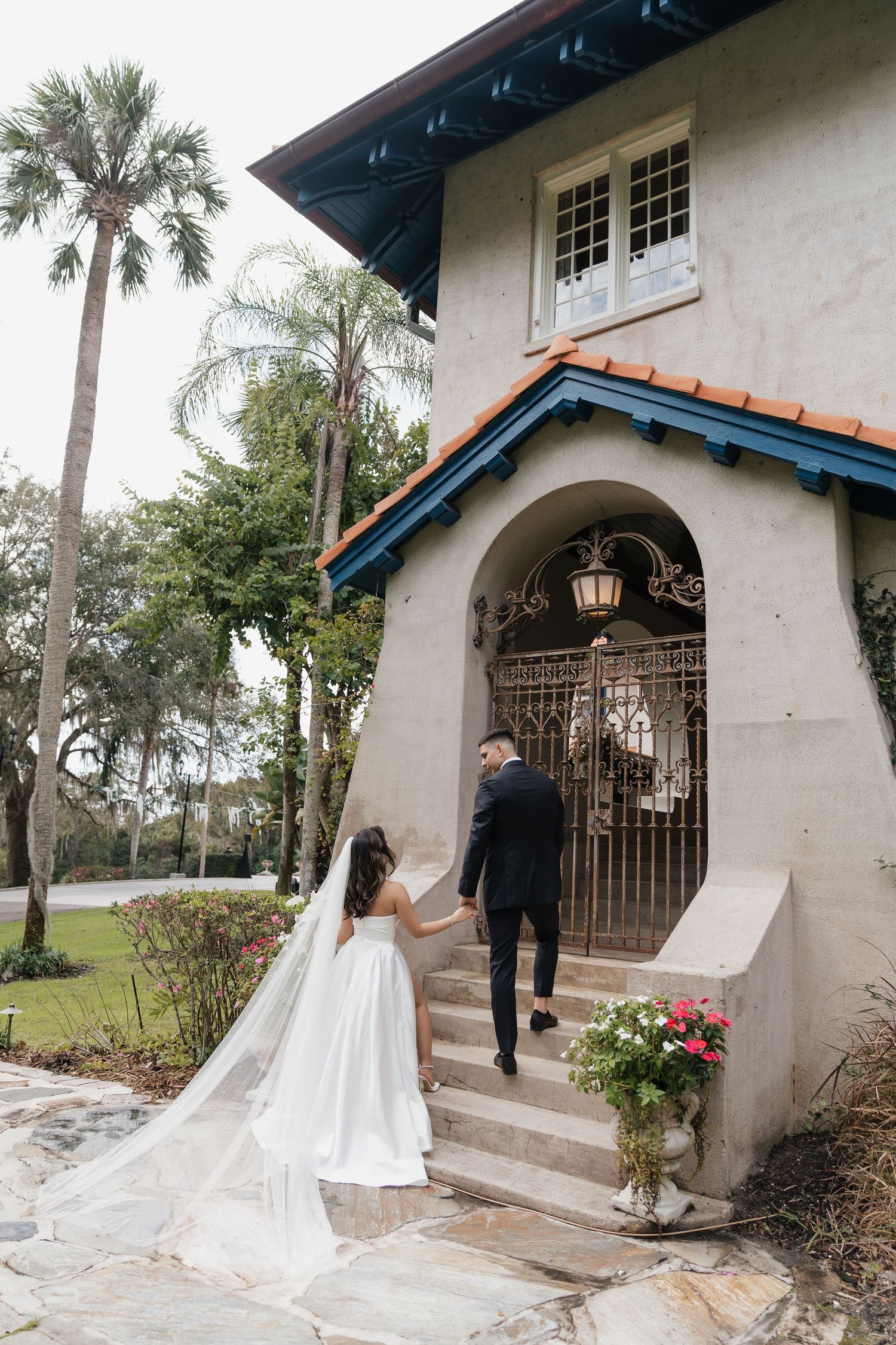 Bride and groom holding hands walking up steps to elegant Spanish-style villa entrance on their wedding day.