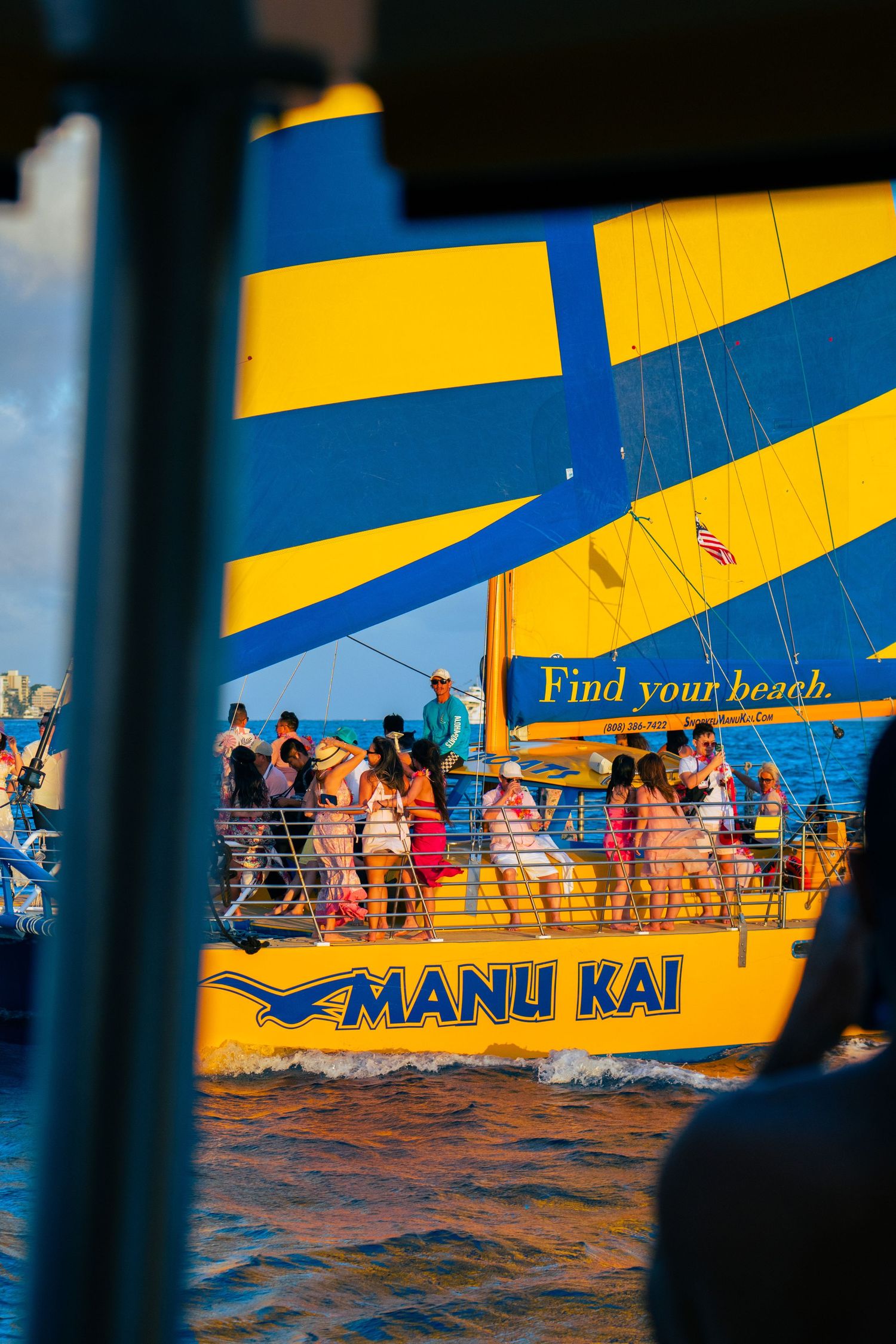 Manu Kai catamaran sailboat with blue and yellow sails cruising with passengers on deck in tropical waters.