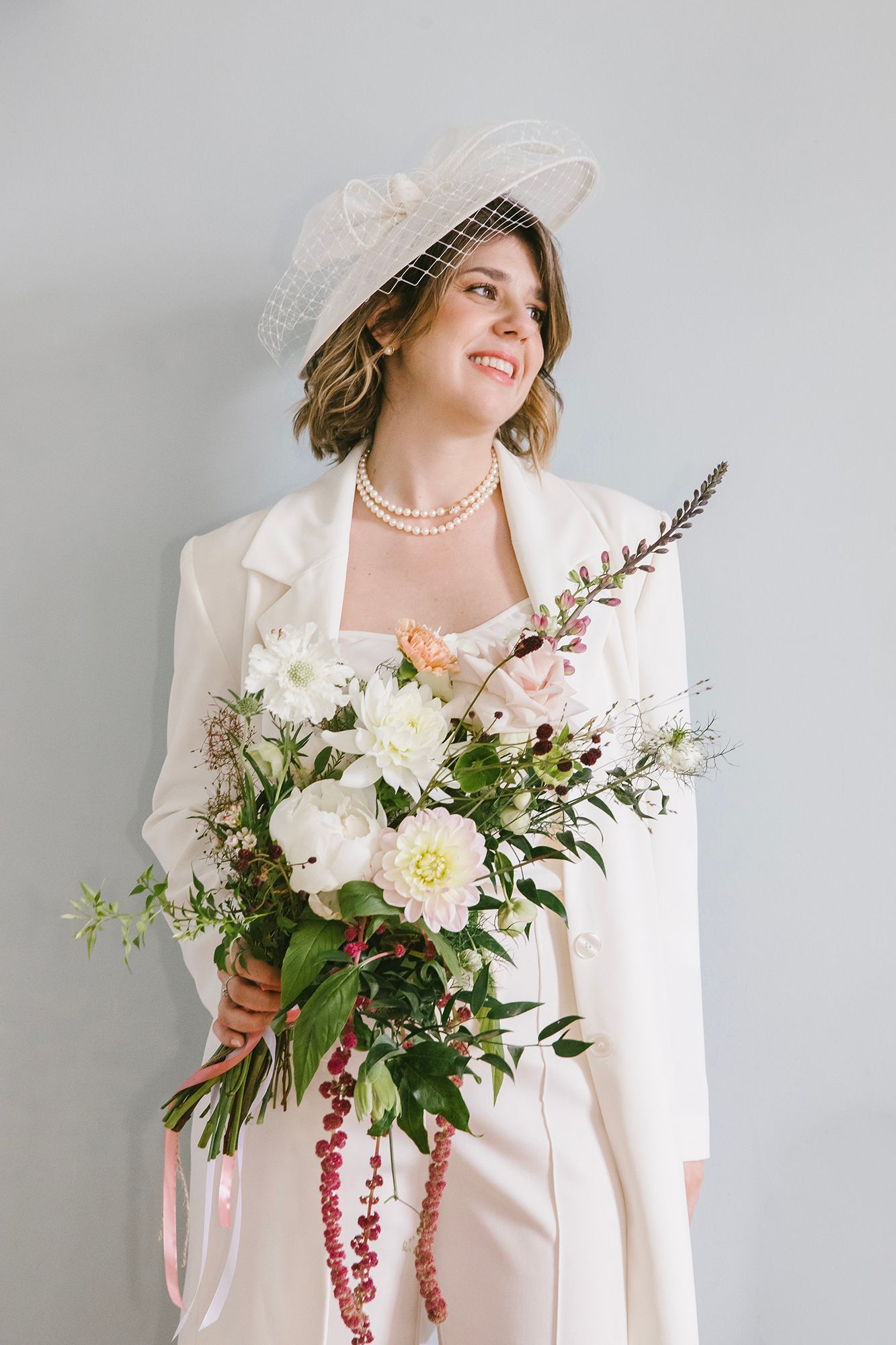 Smiling bride in white coat and veil hat holds a lush bouquet of white dahlias and greenery.