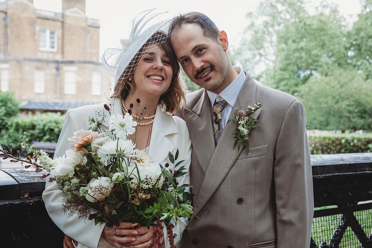 Smiling bride and groom pose outdoors, bride holding white bouquet, groom in tan suit, on their wedding day.
