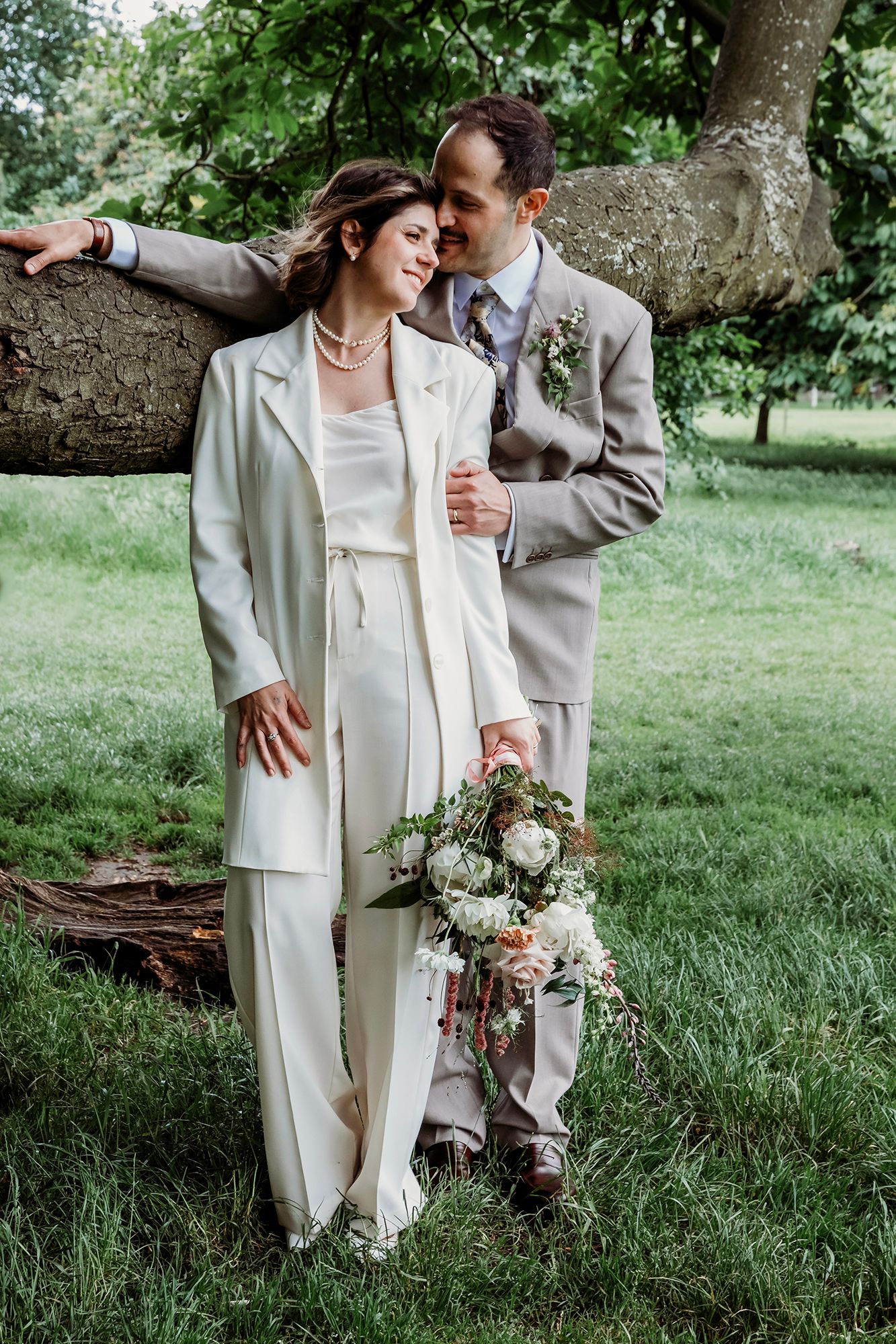 Bride in white pantsuit and groom in gray suit embrace near a tree, holding a floral bouquet outdoors.