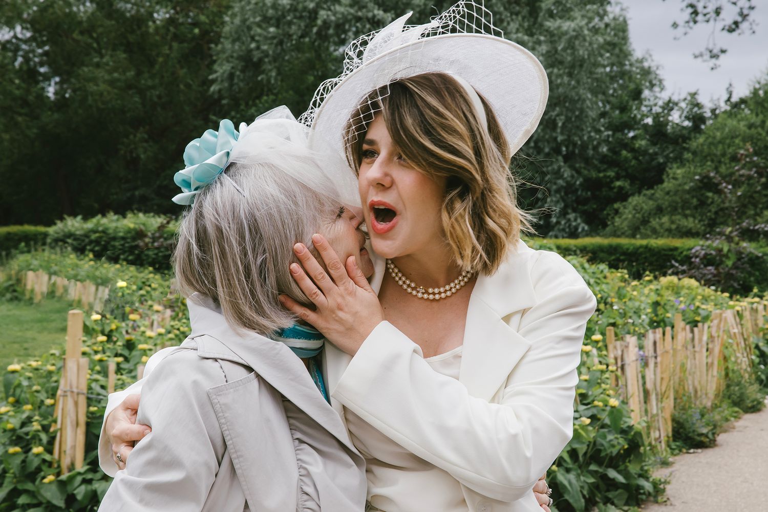 Bride in white hat whispers to older woman with blue flower hair clip at outdoor wedding ceremony.