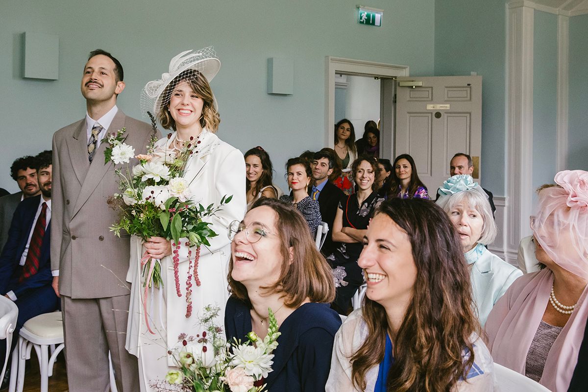 Bride in white suit and hat holds flowers beside groom at a joyful indoor wedding ceremony with smiling guests.