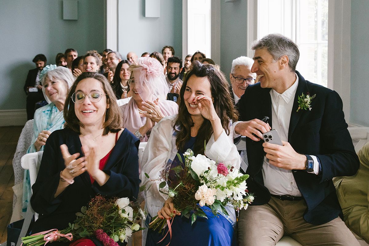 Joyful wedding guests laughing and celebrating during a ceremony, with the bride holding a bouquet of pink and white flowers.