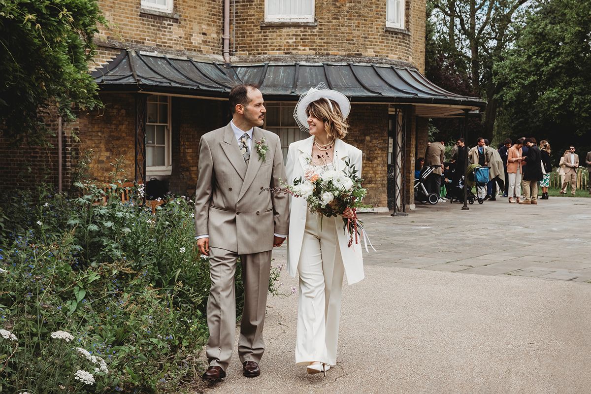Bride in white pantsuit holding bouquet walks with groom in grey suit outside a brick building on their wedding day.