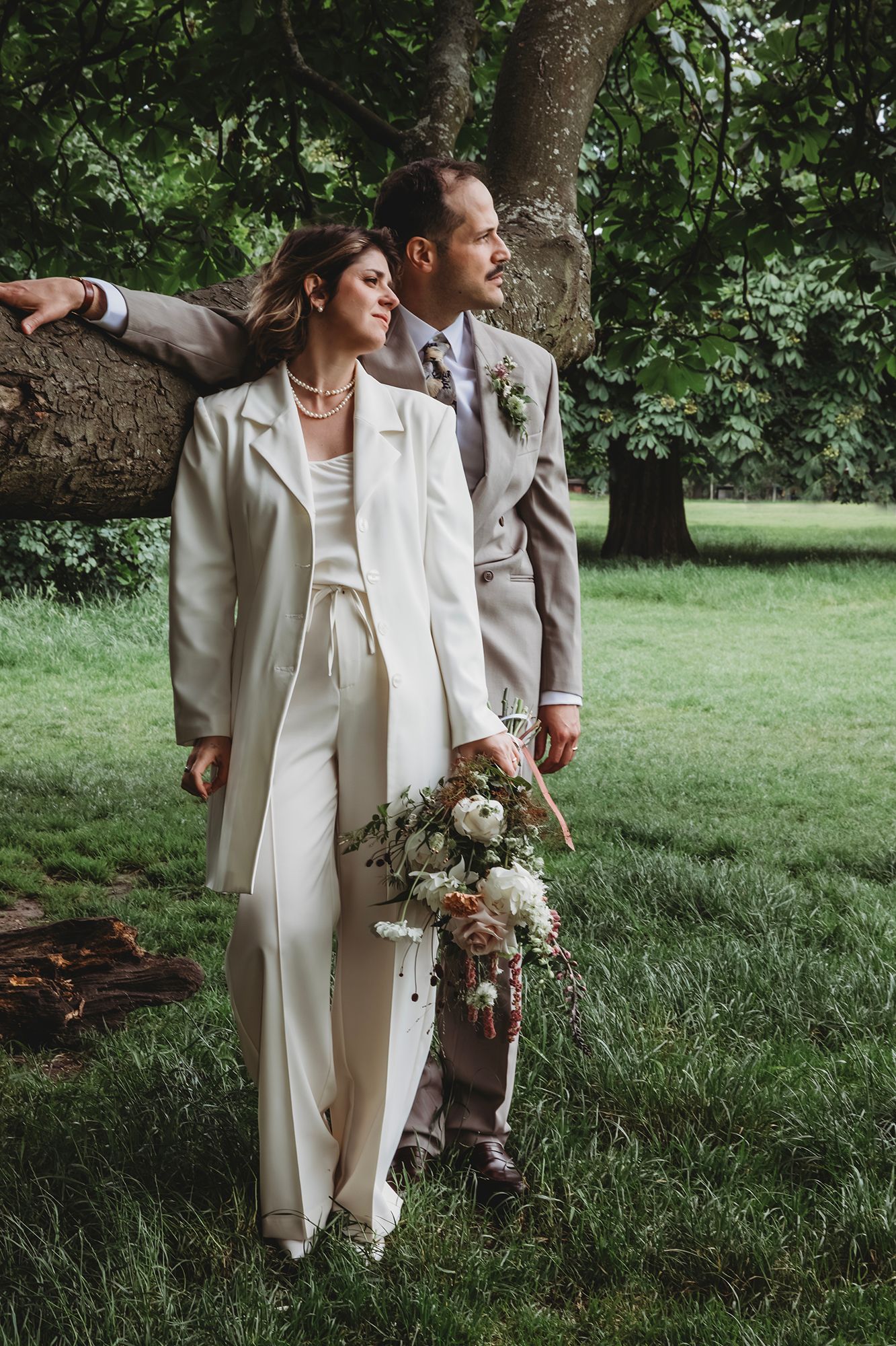 Bride in white pantsuit holding bouquet stands with groom in gray suit outdoors near large tree on lush green lawn.