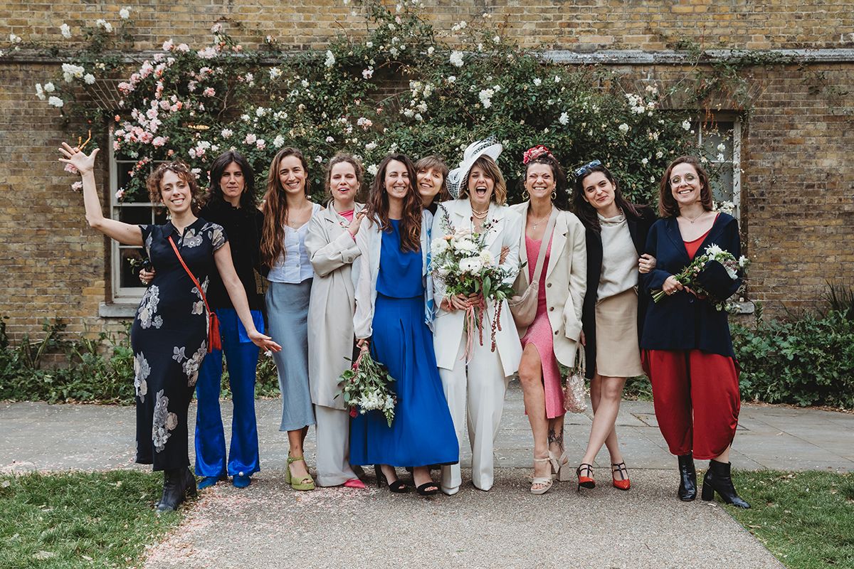 Bride in white suit poses with bridesmaids in colorful outfits in front of flowering wall.