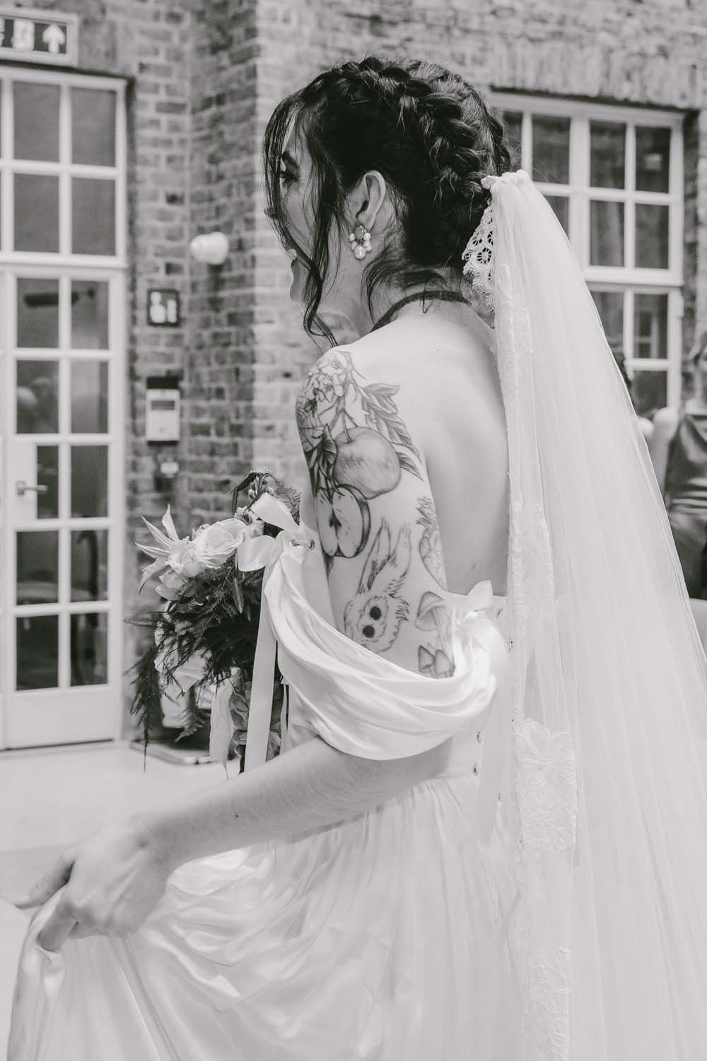 Black and white photo of a bride with back tattoos in a white gown and veil, holding a bouquet outside a brick building.
