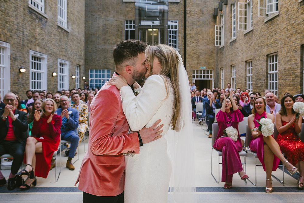 Couple sharing first kiss at outdoor wedding ceremony with cheering guests seated in rows at historic brick venue.