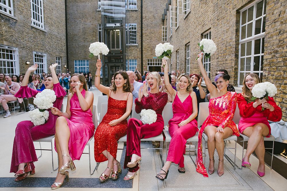 Bridesmaids in pink and red dresses cheering with white bouquets at an outdoor wedding ceremony.