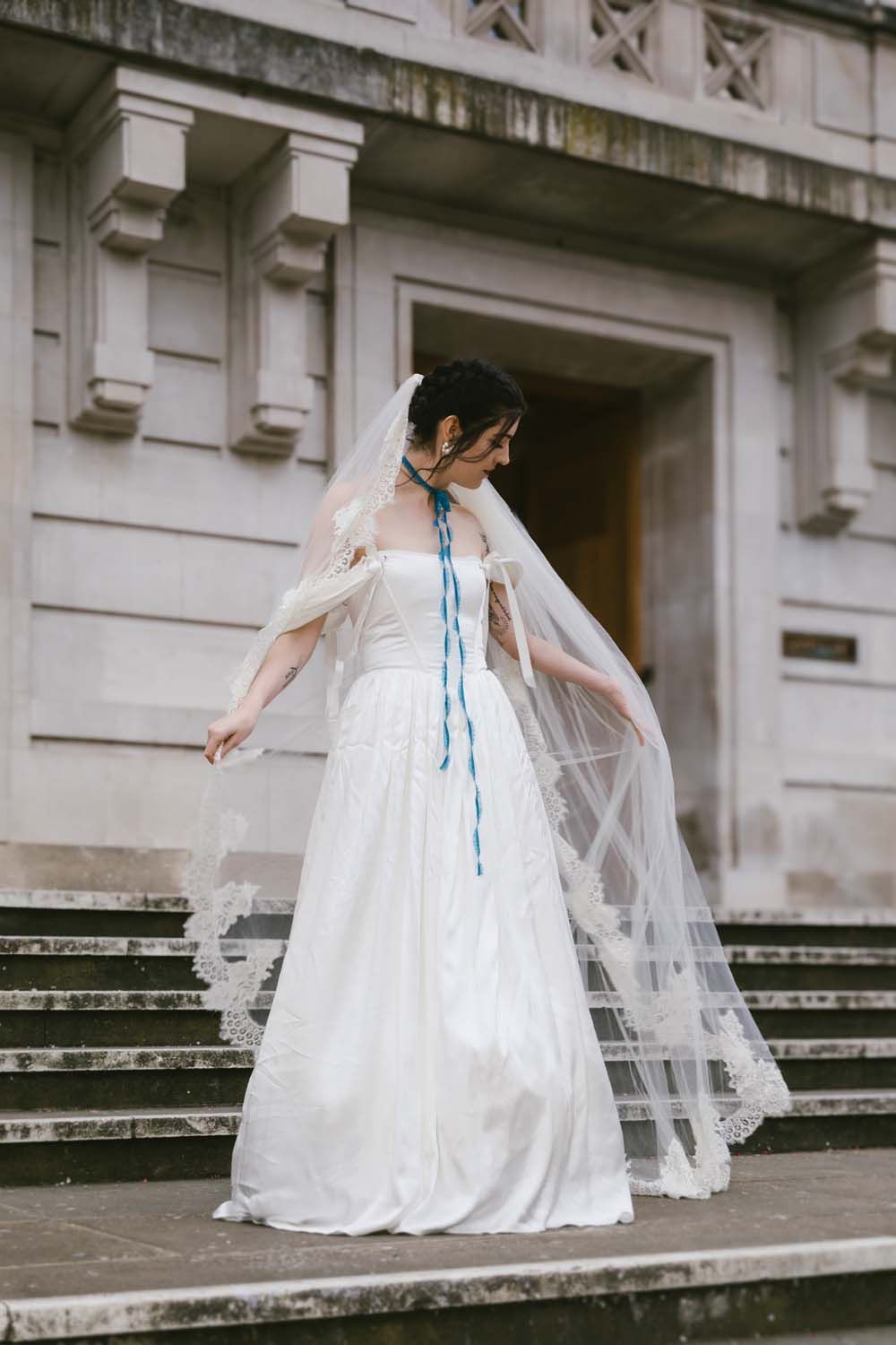 Bride in white gown with blue ribbon veil poses on stone steps of elegant building.