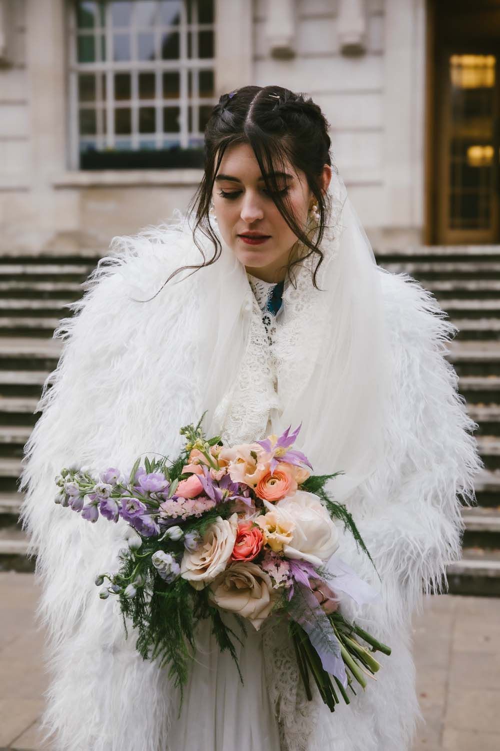 Bride in white lace gown and fluffy white wrap holds colorful spring bouquet on stone steps.
