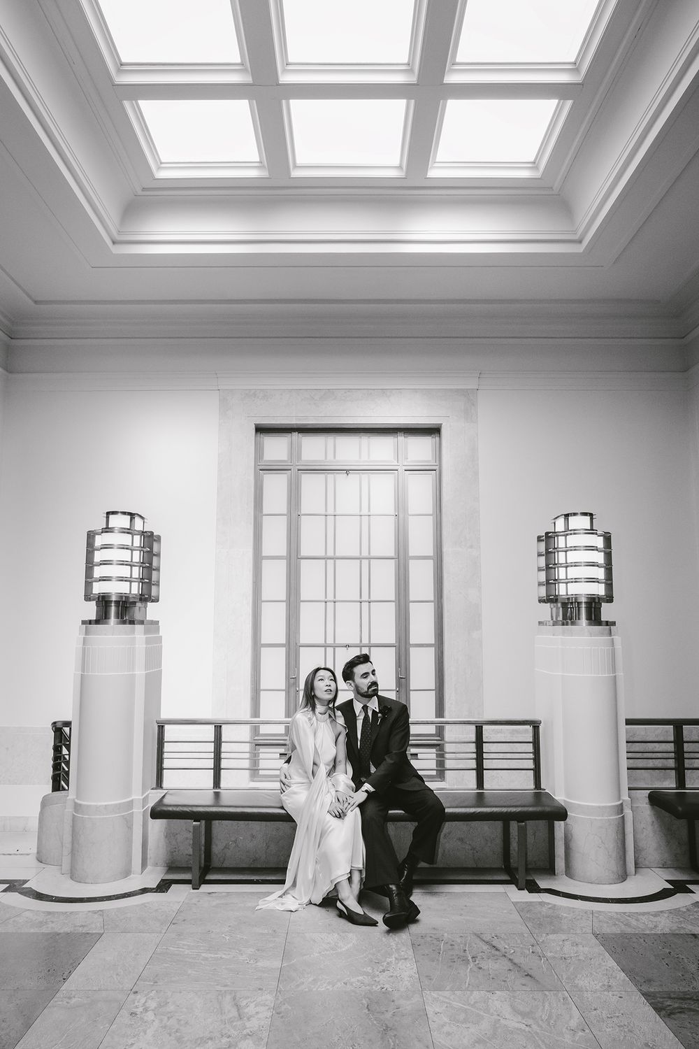 Black and white photo of bride and groom seated on bench flanked by Art Deco lamps beneath skylight windows.