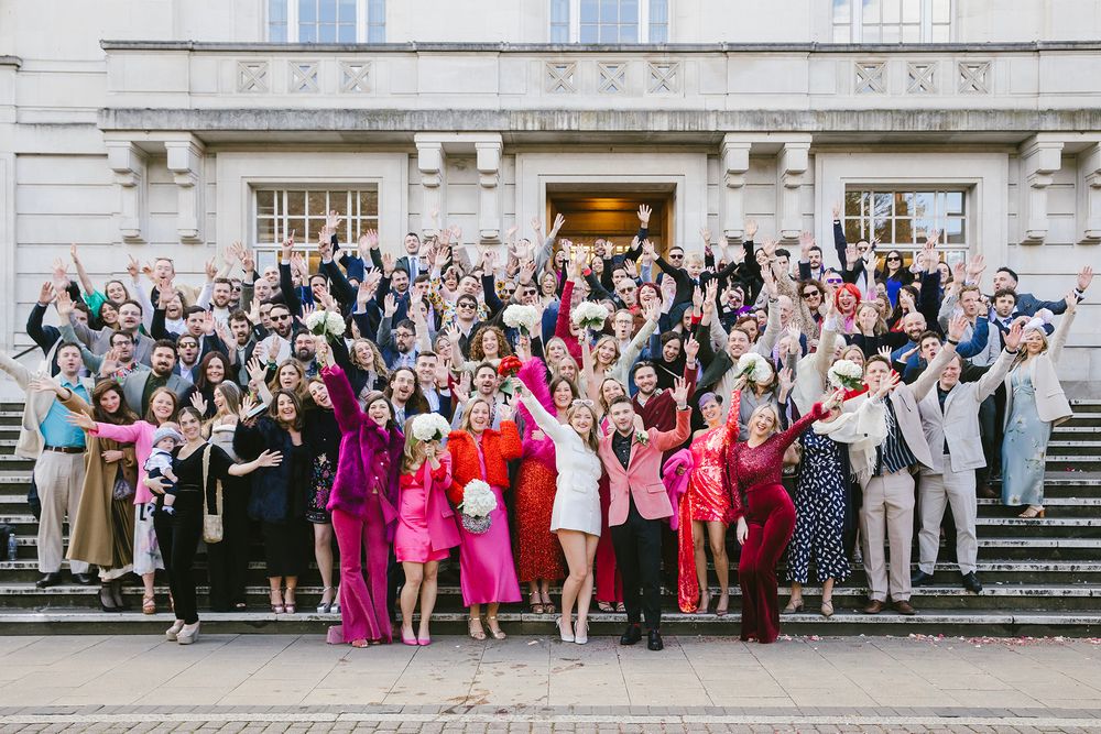 Large wedding group photo on building steps, guests in colorful outfits cheering with arms raised in celebration.