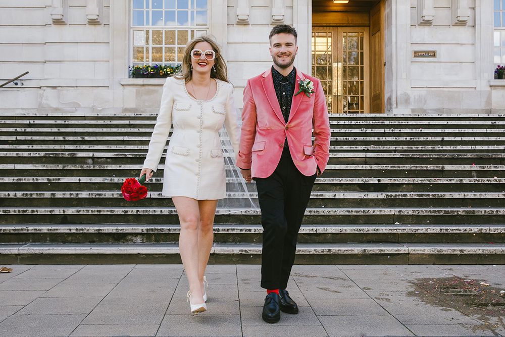 Bride in white mini dress and groom in pink blazer holding red roses on steps of a registry office.