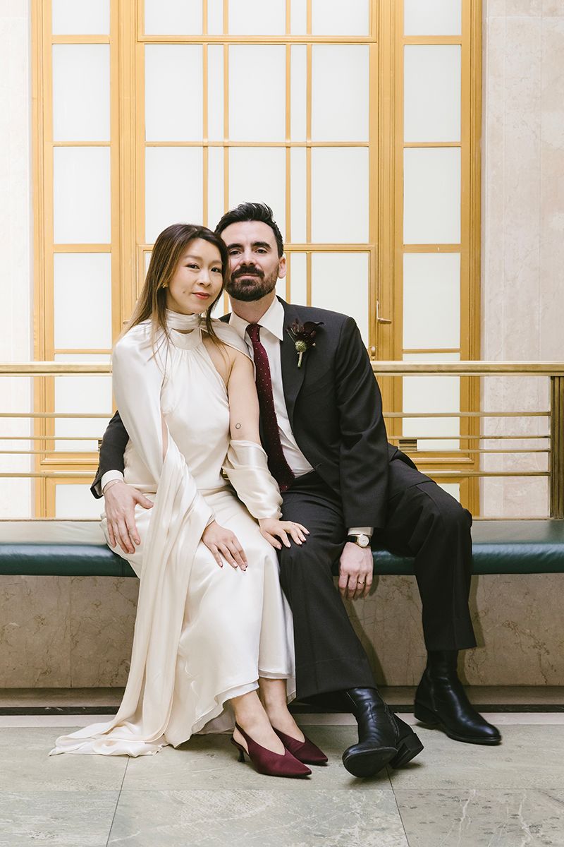 Elegant couple in wedding attire sitting together on a bench, woman in white dress and man in dark suit with burgundy tie.