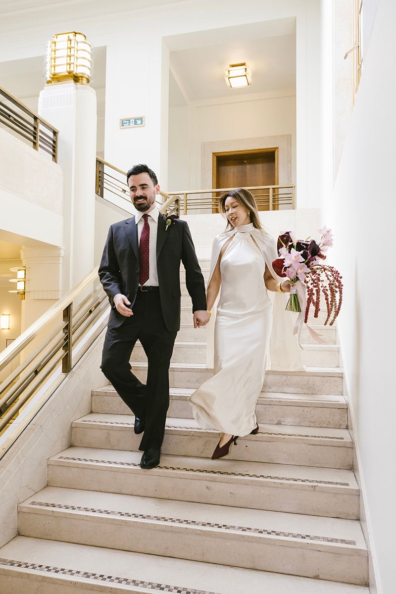 Bride and groom walk hand-in-hand down elegant marble staircase, she in white gown with bouquet, he in dark suit with red tie.