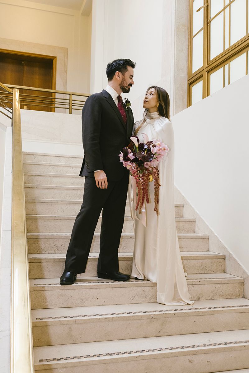 Bride and groom share a romantic moment on marble stairs, she holds a burgundy bouquet in an elegant white gown.