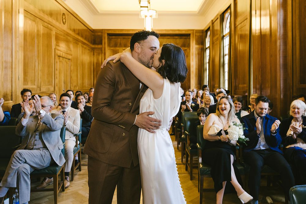 Bride and groom share first kiss at wedding ceremony while guests applaud in elegant wood-paneled room.