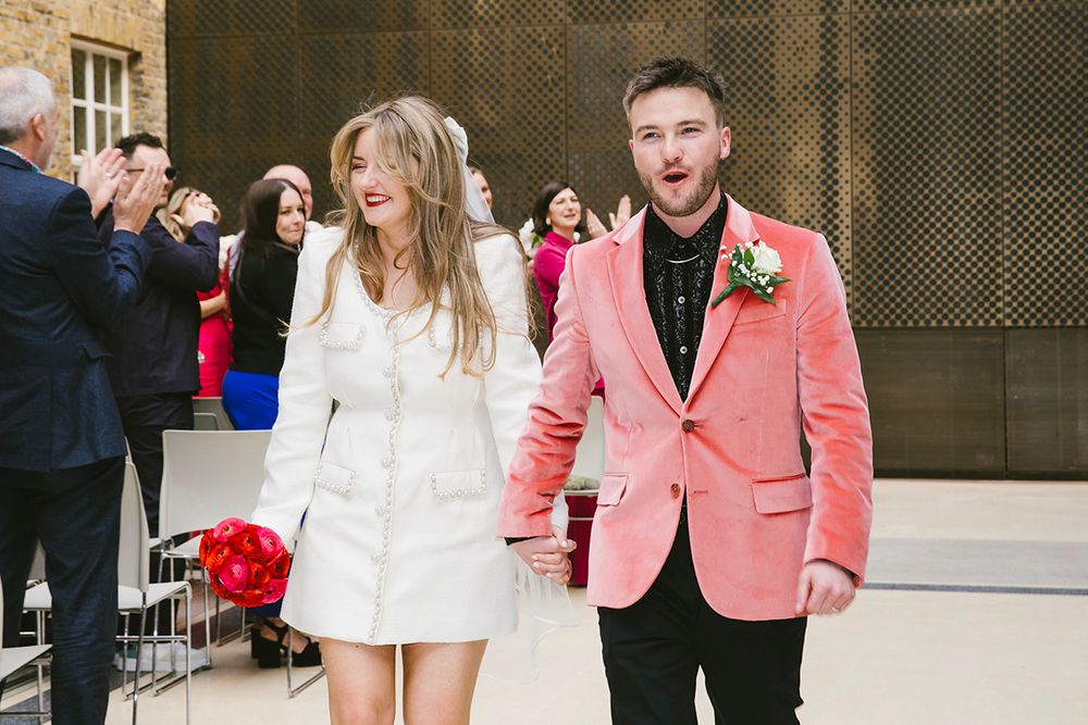 Bride in white mini dress and groom in pink velvet blazer walking together after wedding ceremony.