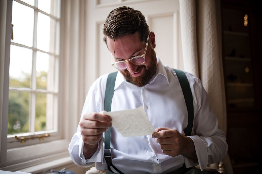 Groom in white shirt and suspenders reading a heartfelt letter by a window on his wedding day.