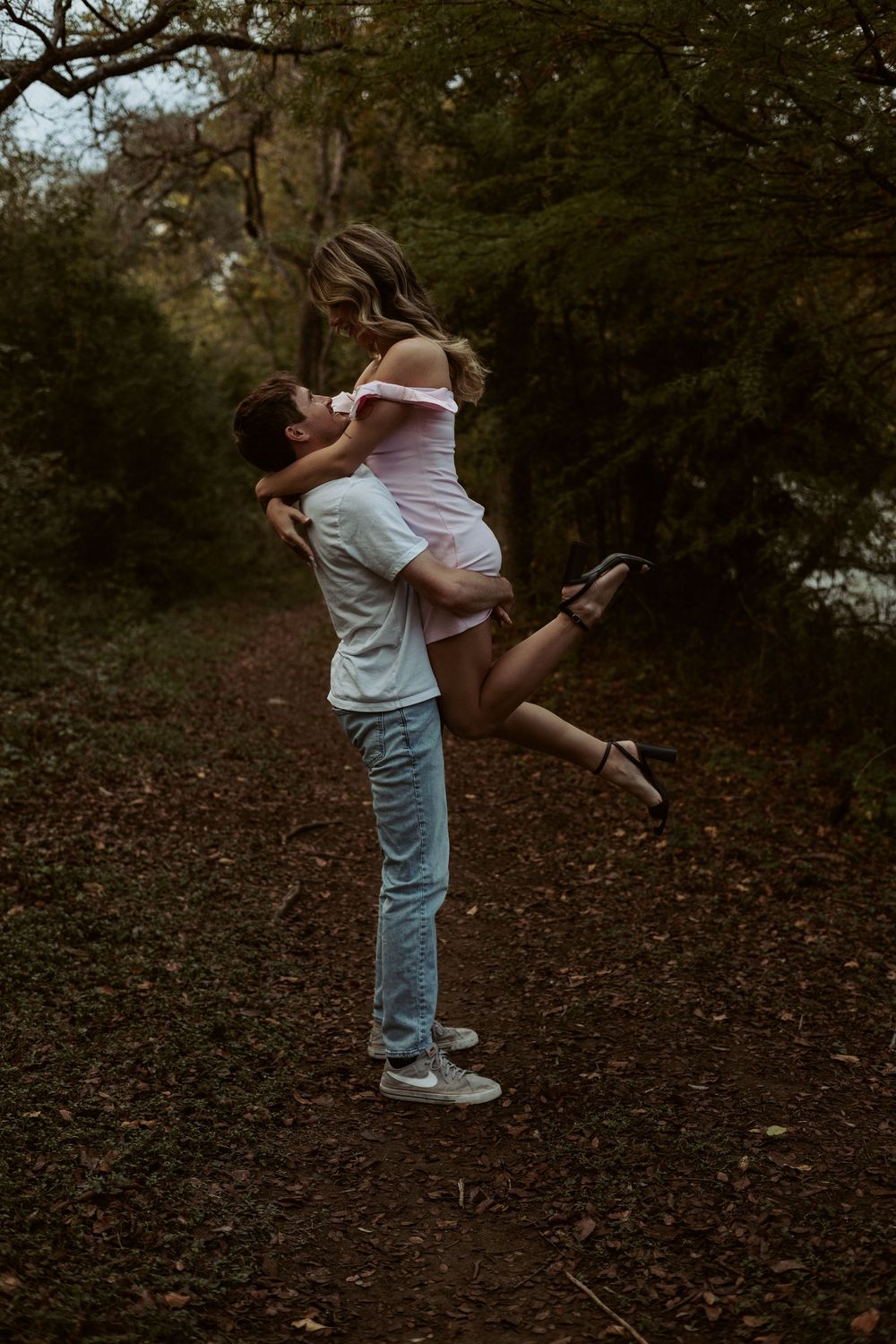 A joyful and candid outdoor lifestyle portrait of a man lifting a woman in a sun-drenched wooded area.