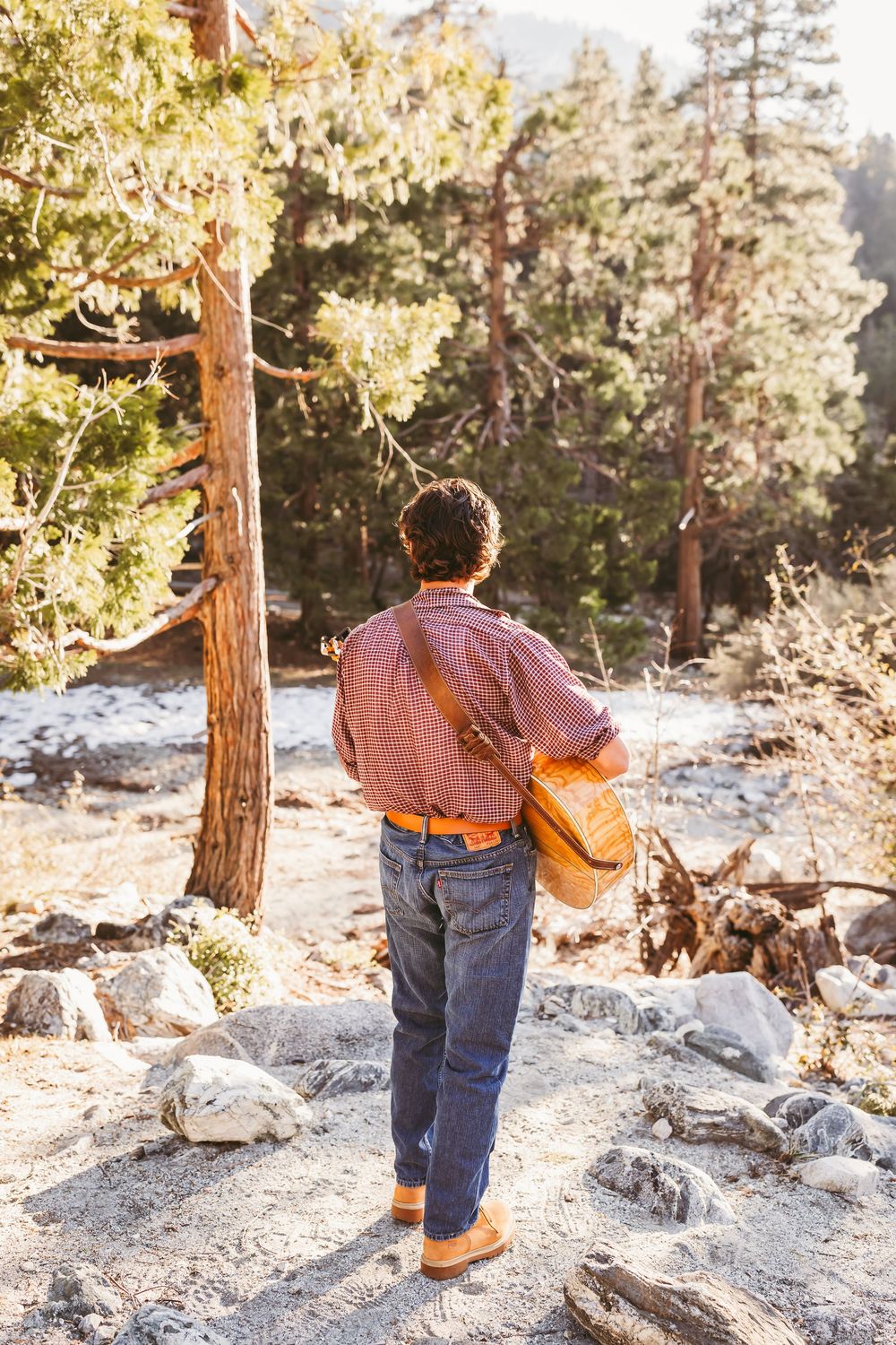 A hiker in a red plaid shirt and jeans carries a yellow bag through a rocky mountain forest trail.