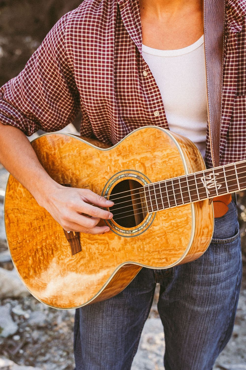 Close-up of a musician strumming an acoustic guitar, wearing a plaid shirt and jeans outdoors.