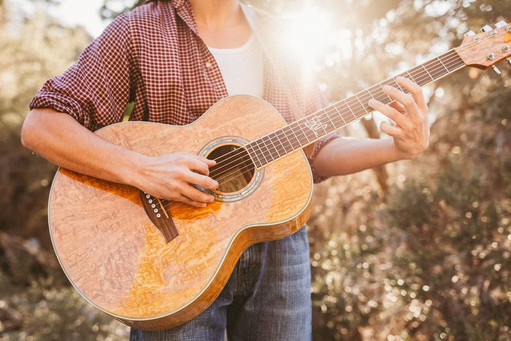 Person playing acoustic guitar outdoors in sunlight wearing red plaid shirt.