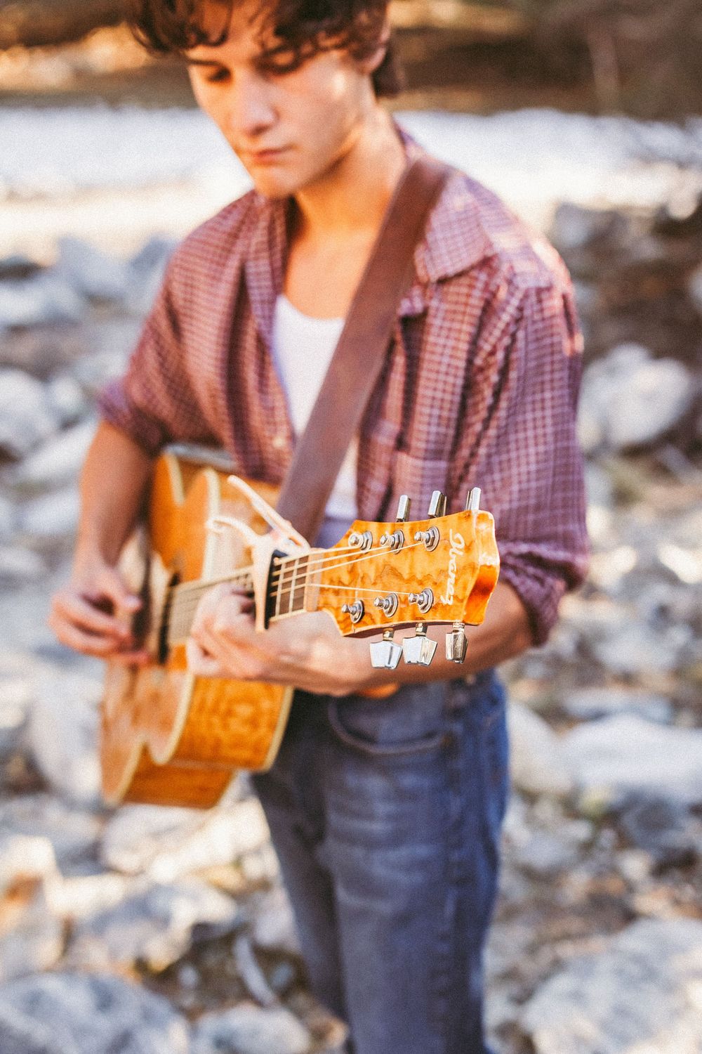 Young man playing acoustic guitar outdoors on rocky terrain, wearing plaid shirt and jeans, focus on guitar headstock.