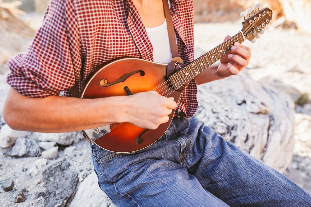Close-up of musician playing a mandolin outdoors, wearing a red plaid shirt and jeans.