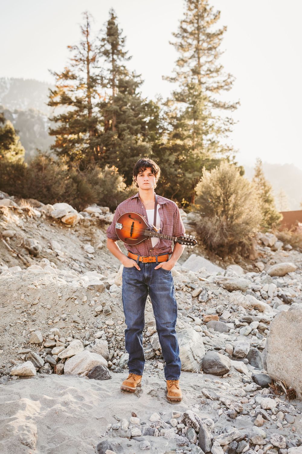 Young man holding a mandolin standing on rocky terrain with pine trees in background at golden hour.